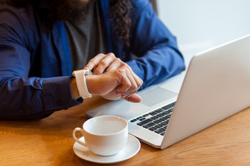 Close up portrait of young adult man freelancer in casual style sitting in cafe with laptop and cup of coffee, looking to the watch to check the time, bussinessman in office. Indoor, lifestyle concept