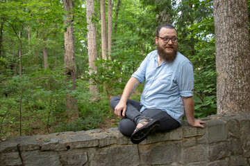 Naklejka na ściany i meble Young bearded man sits on a stone wall with an amused expression, looking away from camera. Relaxed hipster with glasses, beard and ponytail in a natural environment.