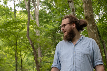 Naklejka na ściany i meble Young man looking around in forest is about to smile. Relaxed hipster with glasses, beard and ponytail in a natural environment.