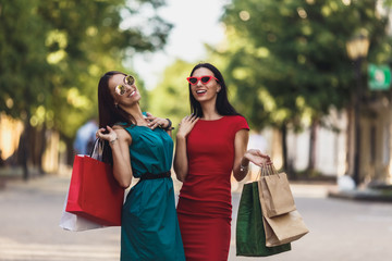 Young attractive girls with shopping bags in the summer city. Beautiful women in sunglasses looking at camera and smiling. Positive emotions and Shopping day concept.