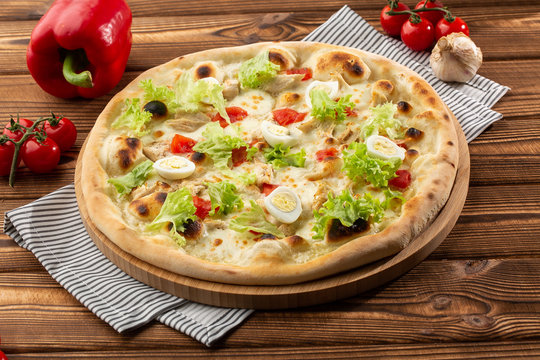 Delicious pizza Caesar style with white sauce, chicken, parmesan, egg, cherry tomatoes and fresh lettuce at wooden background. Restaurant pizzeria menu with delicious taste pizza Caesar 