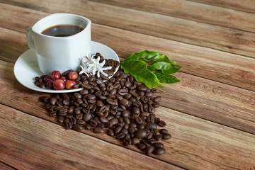 a cup of hot coffee with roasted coffee and leaves, flowers, and coffee fruit on a wooden table
