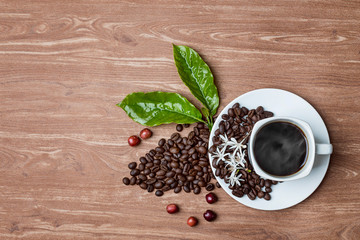 a cup of hot coffee with roasted coffee and leaves, flowers, and coffee fruit on a wooden table