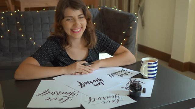 Close up shot of a young woman calligraphy writing on a paper using lettering technique. She looks at sheets with words Love yourself and laughs