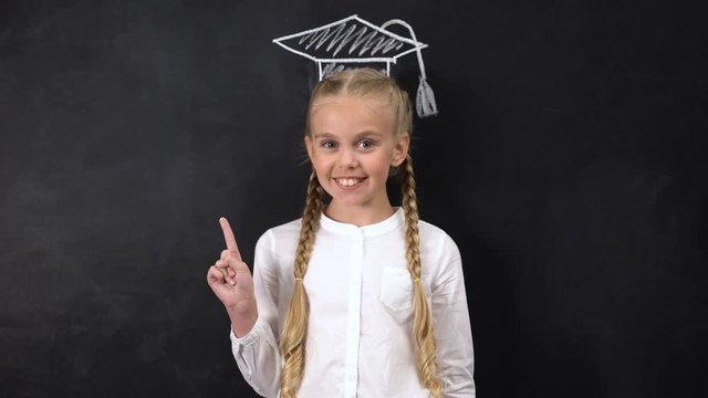 Happy schoolgirl points finger at academic cap painted on blackboard background