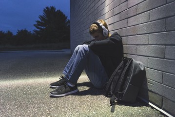 Sad teenage boy sitting on the ground against a brick wall at night. He is listening to music...