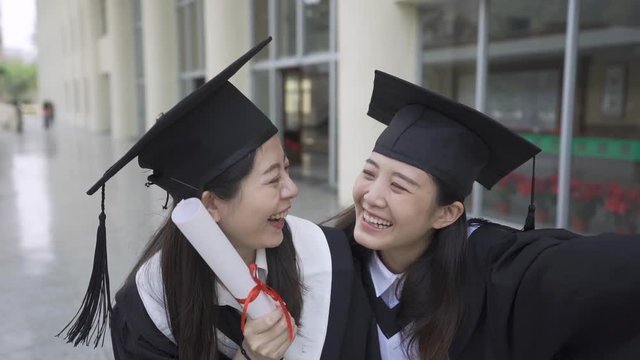 slow action pretty asian girls graduating students taking selfie with diploma scrolls wearing graduation gown and mortarboard in campus building hall. women posing smiling celebrating face camera