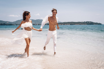 Young beautiful man and woman couple in white clothes running along the beach. Couple man and woman...