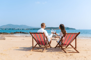 Smiling partners enjoying drinks champagne while resting on comfortable chairs by sea beach. Blue sea on the background. Thailand. Back view