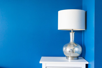 Vibrant blue painted wall with white home decor accents (a white lampshade and silver metallic lamp stand, on a white night table). Room for text and background copy.