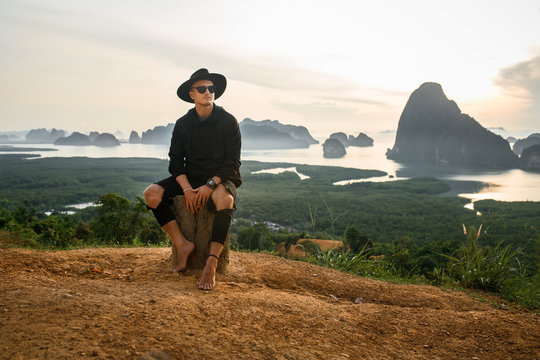 Young casual fashion man  in a hat  and black clothes sitting on a tree stump while looking up.  Incredible sunset background with mountains and lakes. The concept of balance and harmony in life. 