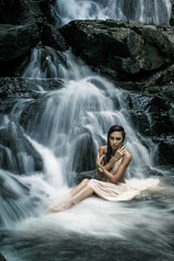 Sensual female in white dress is sitting in the water on a big waterfall.