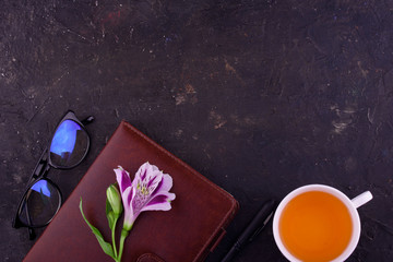 Fragrant tea in a white cup, beautiful flowers, a black background, a notebook with a pen and glasses.