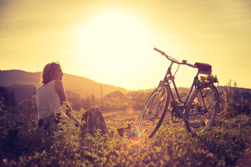 Obraz na płótnie Canvas Enjoying the evening sun, sundown scenery: woman with bike is sitting in the green grass