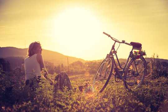 Enjoying the evening sun, sundown scenery: woman with bike is sitting in the green grass