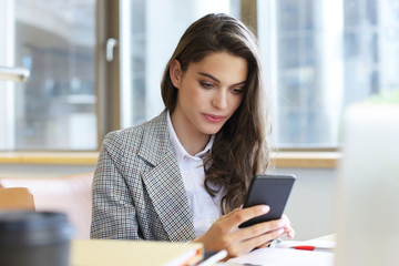 Fototapeta premium Young smiling business woman using smartphone near computer in office.