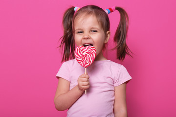 Close up portrait of beautiful female child holding big pink spiral lollipop, looking happy, posing...