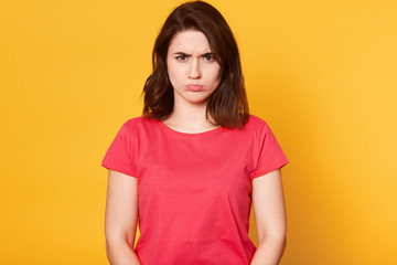Fototapeta premium Portrait of upset miserable black haired woman with frowned face standing isolated over bright yellow background in studio, looking directly at camera, wearing casual red t shirt, being frustrated.
