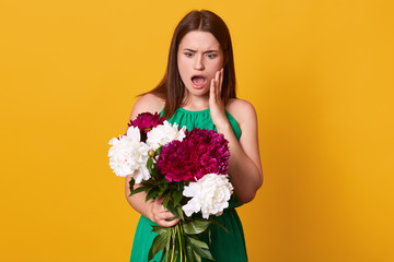 Beautiful girl standing with bouquet of burgundy and white peonies in her hands, wearing green sundress, posing with open mouth, has astonished facial expression, isolated over yellow background.