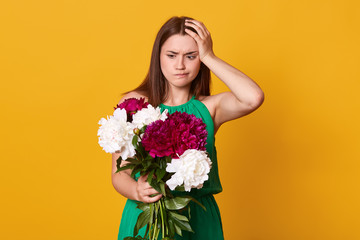 Studio shot of beautiful brunette girl holds big bouquet of burgundy peonies in hand, thoughtful woman with flowers keeps hand on head, poses against yellow studio background. Copy space for promotion