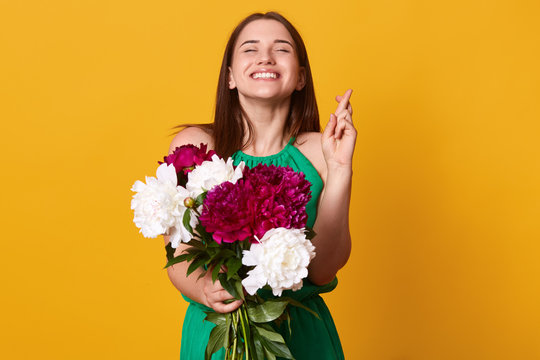 Stidio shot of pretty Caucasion woman raises crossed fingers, prays for good luck, has pleased and happy expression, wears green sundressr, holds white and burgundy peonies, believes in fortune.