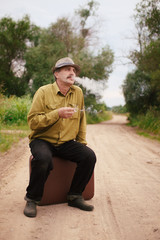 An elderly man travels with a suitcase. A villager is on the road to a long way and smoking a cigarette.