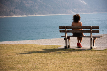 Naklejka na ściany i meble A girl sitting over her shoulder on a wooden bench looks at a mountain lake. The girl with her back to him looks naked, wearing a black skirt and red shoes with high heels. Concept of feminine sensual