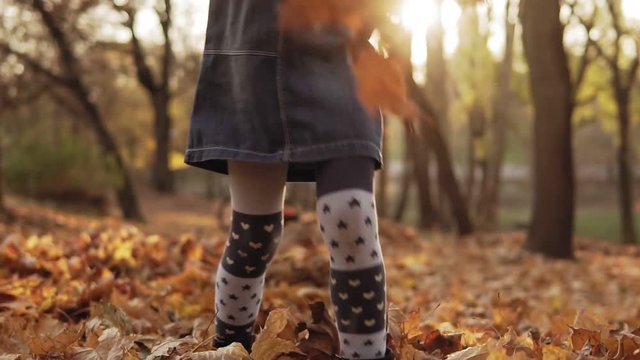 Close up of little girl kicking autumn leaves by boots in park. Slow motion. Low angle. View of legs