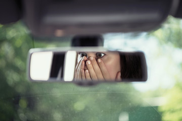 Reflection of young woman with frightened eyes covering her face with  hands in the car rear view...