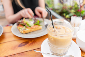 Focus in foreground on Cold coffee with ice cubes. A young woman is having Breakfast in a summer cafe.