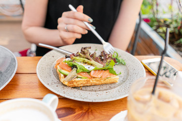 Traditional Belgian waffle with salmon, lettuce leaves and poached egg. A young woman is having Breakfast in a summer cafe, hands with a fork and a knife close-up. Cold coffee with ice cubes