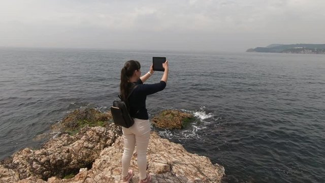 A girl with a tablet in her hands stands on a rocky beach and takes pictures of a beautiful view of the camera