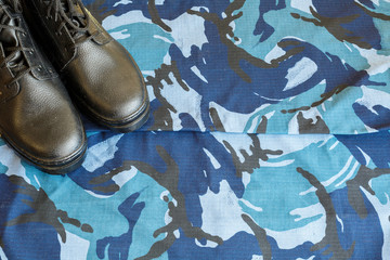 A pair of black army boots on fabric with blue camouflage with copispace for text.