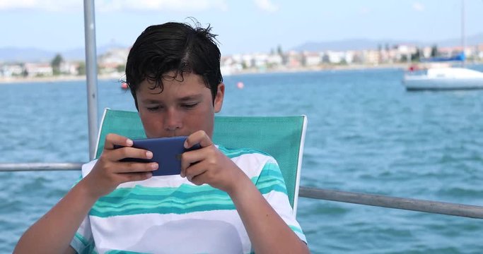 Portrait of a happy caucasian teenager playing video game with smartphone on yacht deck at sunny summer vacation day