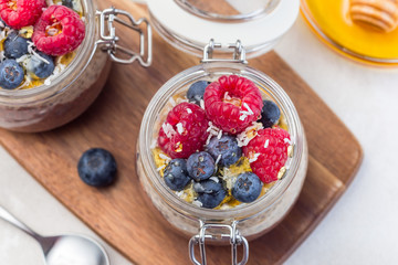 Layered chocolate and peanut butter chia seed pudding in jar, garnished with raspberry, blueberry, honey and coconut flakes, horizontal, top view