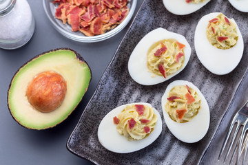 Deviled eggs stuffed with avocado, egg yolk and mayonnaise filling, garnished with bacon,...