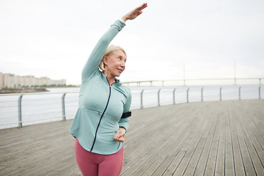Mature active female in sportswear doing side-bends while training by riverside in urban environment on summer morning