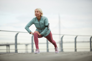 Mature sportswoman doing exercise for stretching legs while working out on bridge in urban environment on summer day