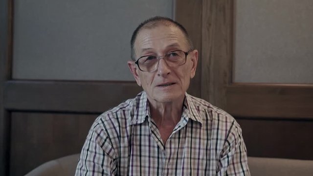 Old man sitting on the porch of the house. Pensioner turns face and looking to the camera, smiling, talking.
