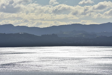 Sea surface with silver reflection from back light and hilly coast in background.