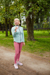 Mature healthy active female with bottle of water standing on path in city park among green trees after morning workout