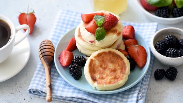 Syrniki or cottage cheese pancakes with fresh strawberries, blackberries, honey and cup of black coffee on side. Footage of tasty healthy breakfast