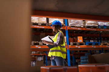 Warehouse worker walking through large storage area checking inventory and organizing goods distribution. Smiling woman employee working in warehouse distribution center.