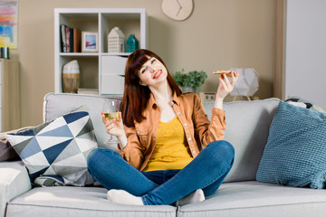 Happy young smiling girl in jeans and yellow shirt, holding glass with wine and piece of pizza , while sitting on gray sofa with pillows at home