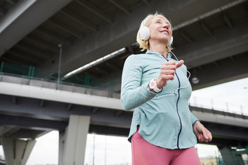 Happy aged blonde active female in sportswear and headphones enjoying music while jogging in urban...