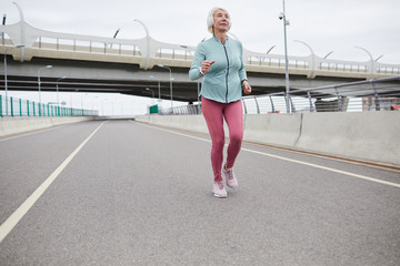 Contemporary mature female in activewear listening to music in headphones while running along road in the city