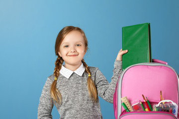 Closeup portrait of little girl schoolgirl on blue background. The child puts in a satchel book....