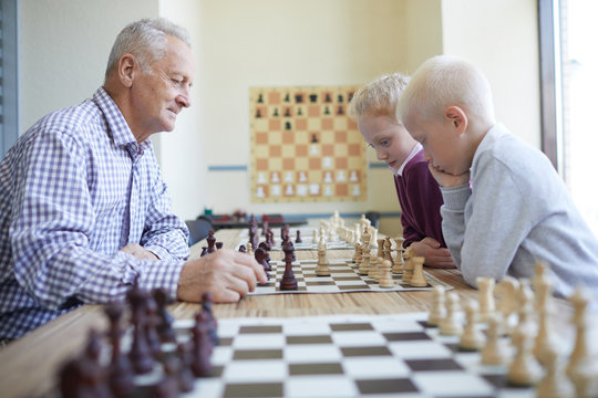 Old man in checked shirt playing chess with two focused schoolchildren studying at chess club