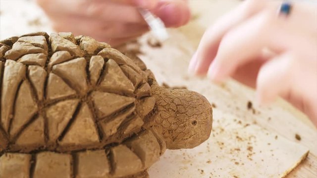 Creating toy made of clay in the workshop. Child is scratching with spicial tool the texture of the scales on the handmade turtle toy's head from clay, hand closeup.