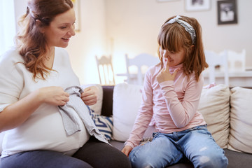 Happy pregnant Caucasian mom sitting with her cute daughter on sofa in living room and holding...